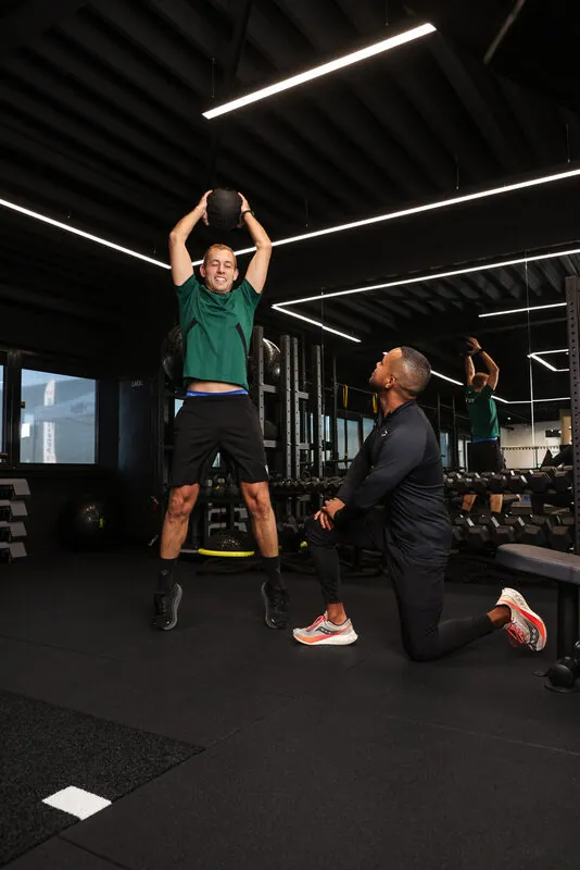 Klant tilt een medicine ball boven het hoofd, trainer kijkt de uitvoering na